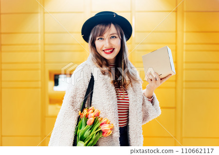 Portrait of smiling fashion woman with box near modern postal automatic mail terminal with self service device for pickup or refund an order. Electronic locker for storing parcels. Selective focus 110662117