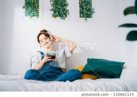 Woman with closed eyes listen music and enjoy cup of coffee or tea. Calm female spend free time at home enjoy favorite song with wireless modern headphones. Pastime weekend relax, no stress concept. 110662124