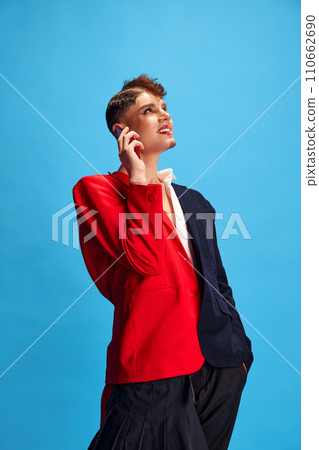 Duet of masculine and feminine voices. Young attractive male model with female and male make-up talking on phone against blue background. 110662690