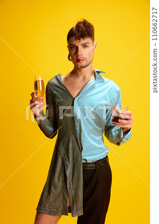 Portrait of person half-wearing in woman's and man's clothes posing holding glasses of champagne and whiskey in hands on his female and male sides. 110662717