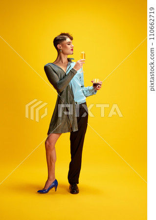 Young male model half-wearing in woman's and man's clothes posing holding glasses of champagne and whiskey in hands on his female and male sides. 110662719