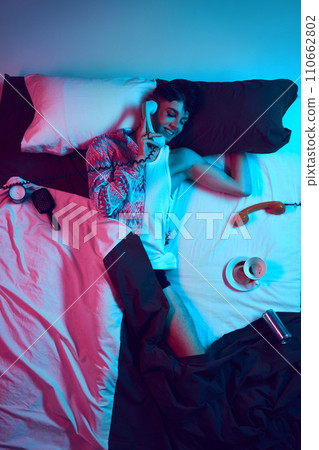 Gender fusion symphony. Visual overture. Creative portrait of young male model lying in bad and talking on retro phone as woman in neon light. 110662802