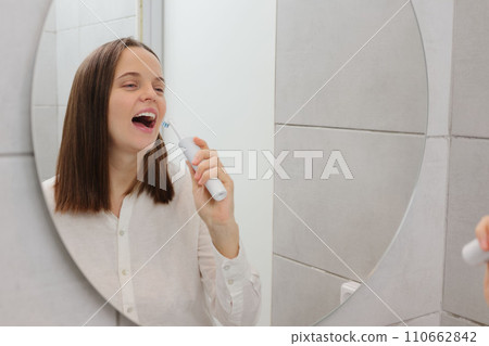 Personal care essentials. Dentistry in the morning. Optimal teeth protection. Caucasian young adult woman brushing teeth in bathroom having fun singing pretending holding microphone in front of mirror 110662842