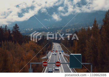 A Highway Through Fall Foliage with Mountains and Cloudy Skies in the Background A Highway Through Fall Foliage with Mountains and Cloudy Skies in the Background 110662886