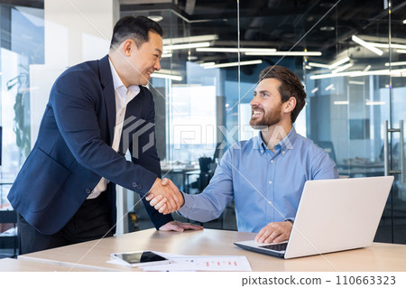 Two professional businessmen in a modern office setting engaging in a friendly handshake, symbolizing a successful agreement or partnership. 110663323