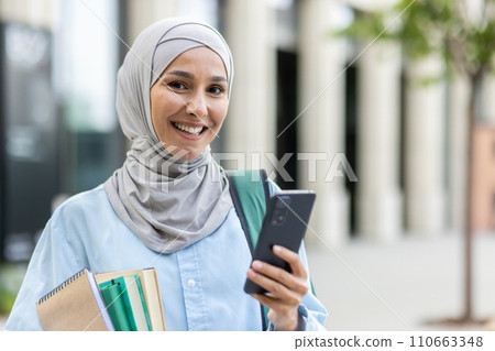 Smiling young Arab woman in hijab holding notebooks and a smartphone outside an office, symbolizing empowerment and education. 110663348