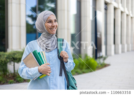 A cheerful young Arab woman in a hijab holding notebooks, walking by an office, exuding confidence and professionalism. 110663358