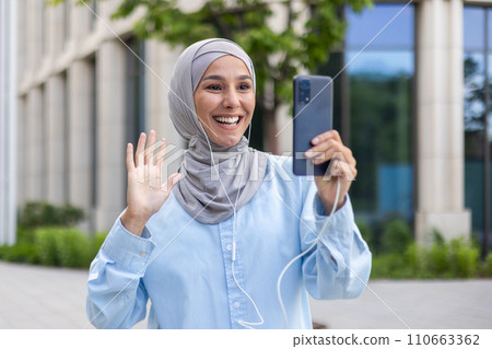 Smiling Arab businesswoman in hijab waving during a video call. Young female student or office worker outside modern building. Smiling Arab businesswoman in hijab waving during a video call. Young female student or office worker outside modern building. 110663362