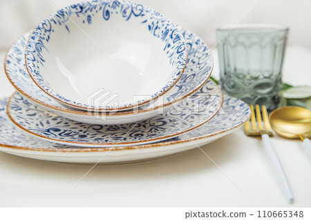 A set of plates of different sizes with a white middle and blue patterns on the side of the plate, stand on top of each other with a tilt, a fork and a spoon of gold - white color are located on the A set of plates of different sizes with a white middle and blue patterns on the side of the plate, stand on top of each other with a tilt, a fork and a spoon of gold - white color are located on the 110665348