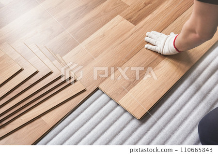 Installing laminated floor, detail on man hand in white glove fitting wooden tile, over white foam base layer Installing laminated floor, detail on man hand in white glove fitting wooden tile, over white foam base layer 110665843