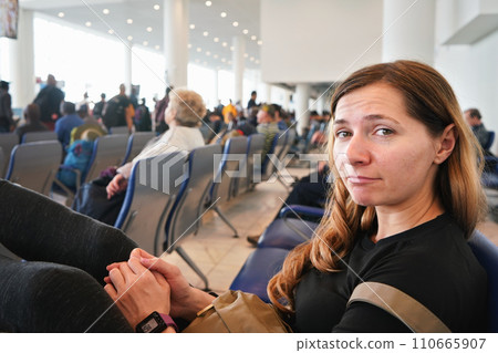 Young woman sitting at airport hall, looking tired after waiting couple of hours for connecting flight in early morning, more blurred passengers in background Young woman sitting at airport hall, looking tired after waiting couple of hours for connecting flight in early morning, more blurred passengers in background 110665907