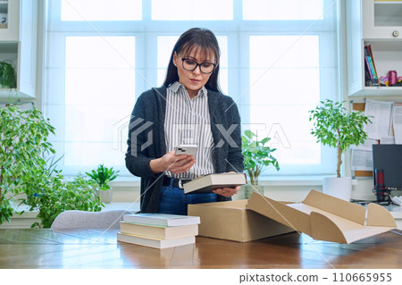 Woman with smartphone unpacking box with books, online shopping 110665955