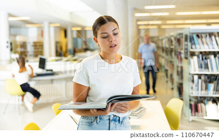 Carefree young lady in everyday clothes reading the magazine and smiling while standing in library during daytime 110670447