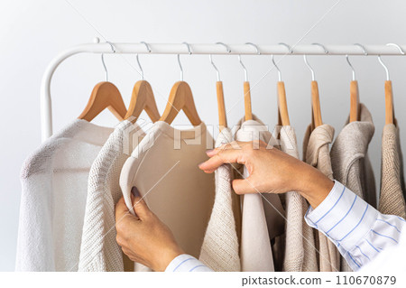 Women's hands choose neutral clothes from a hanger. White, beige and gray jersey. Women's hands choose neutral clothes from a hanger. White, beige and gray jersey. 110670879