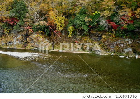 The flow of the Arakawa river and the Chichibu Red Cliffs with autumn leaves above Nagatoro Iwatatami 110671380