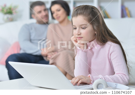 Happy girl sitting at table with laptop with her parents Happy girl sitting at table with laptop with her parents 110674107