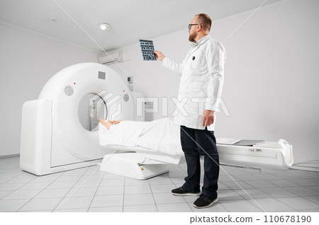 Medical computed tomography or MRI scanner. Doctor holding and examining results of MRI. Woman patient lying on CT scan bed. Concept of medicine, healthcare and modern diagnostics. Medical computed tomography or MRI scanner. Doctor holding and examining results of MRI. Woman patient lying on CT scan bed. Concept of medicine, healthcare and modern diagnostics. 110678190