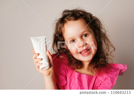 Child little child girl holding white cosmetic cream tube and smiling. Daily children beauty product 110683213