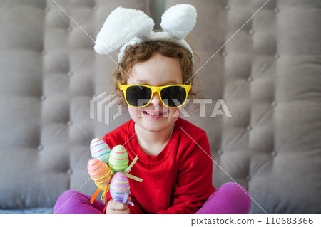 Happy Easter. cute beautiful girl with curly hair, sunglasses and bunny ears sitting on the sofa with colorful easter eggs. Lifestyle photo Happy Easter. cute beautiful girl with curly hair, sunglasses and bunny ears sitting on the sofa with colorful easter eggs. Lifestyle photo 110683366