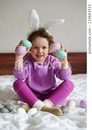 Happy Easter. cute beautiful girl with curly hair and bunny ears sitting on the bed with colorful easter eggs. Lifestyle photo 110683415