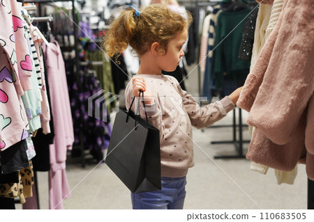 Cute child girl with black paper bag chooses fur coat in shopping mall with clothes Cute child girl with black paper bag chooses fur coat in shopping mall with clothes 110683505