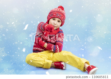 Child in warm clothes on a blue winter background. A boy in a red knitted hat and sweater. Fashionable boy in the studio in white snowflakes Child in warm clothes on a blue winter background. A boy in a red knitted hat and sweater. Fashionable boy in the studio in white snowflakes 110685811