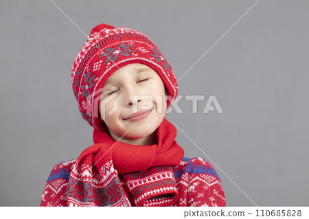 Dreamy child in warm clothes on a gray background. Boy in a red knitted hat with closed eyes. Fashionable boy in the studio on a gray background. 110685828