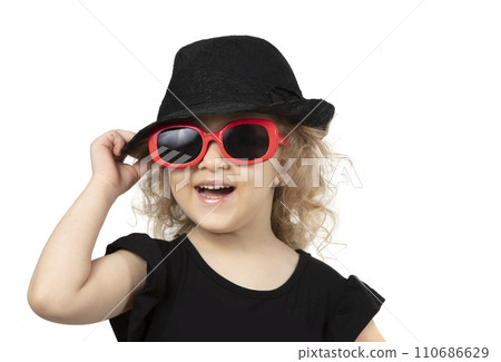 Funny child. Little girl in a black hat and sunglasses on a white background. Portrait of a beautiful fashionable baby girl. Four year old baby blonde. Funny child. Little girl in a black hat and sunglasses on a white background. Portrait of a beautiful fashionable baby girl. Four year old baby blonde. 110686629