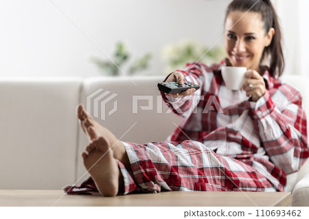 Young brunette woman relaxing on sofa at home watching tv and drinking coffee Young brunette woman relaxing on sofa at home watching tv and drinking coffee 110693462
