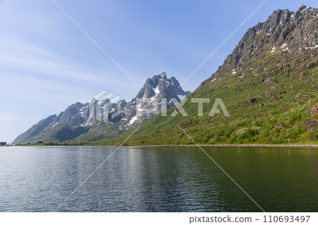 Tranquil Lofoten waters mirror towering peaks with winter remnants under Norway's sky 110693497