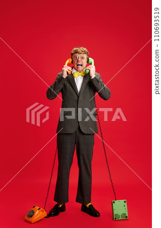 Full-length image of angry, emotional young man, employee, assistant standing with two phones against red studio background 110693519