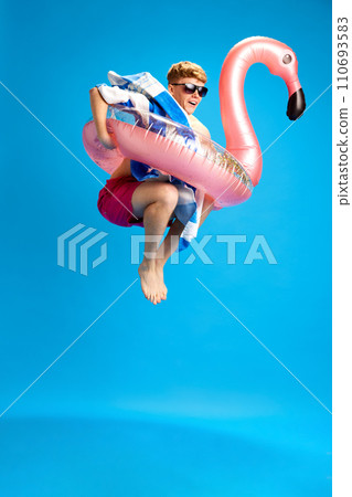 Young cheerful relaxed guy in swimming circle enjoying summer trip, having fun against blue studio background Young cheerful relaxed guy in swimming circle enjoying summer trip, having fun against blue studio background 110693583