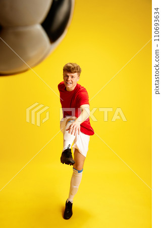 Dynamic image of young man in soccer sportswear, uniform training, hitting ball against yellow studio background 110693634