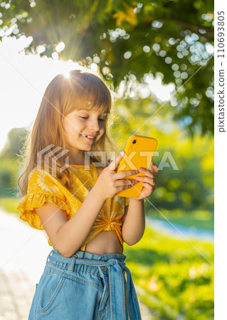Smiling child girl using smartphone typing text answering messages chatting online in city street Smiling child girl using smartphone typing text answering messages chatting online in city street 110693805