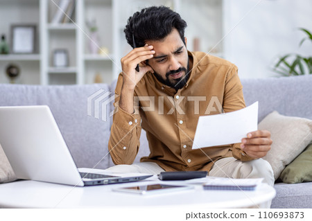 Concerned young Indian man sitting on a couch in home setting, examining papers with a laptop and calculator nearby. Concerned young Indian man sitting on a couch in home setting, examining papers with a laptop and calculator nearby. 110693873