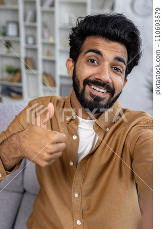 A pleasant young Indian man with a beard smiles and shows thumbs up, seated comfortably on a couch in a well-furnished apartment. 110693879