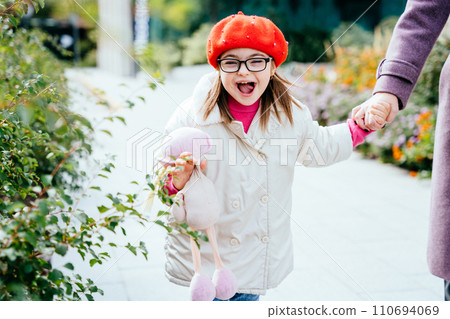 Happy laughing little girl in eyeglasses with special needs holding mother's hand on a walk in autumn time outdoor. Happy family moments concept. 110694069