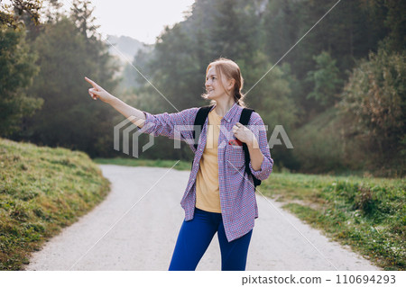 Blonde 30s girl pointing finger on forest background. Young female with backpack hiking on the road in nature. Beautiful woman walking in forest in sport clothes. Freedom and active lifestyle concept 110694293