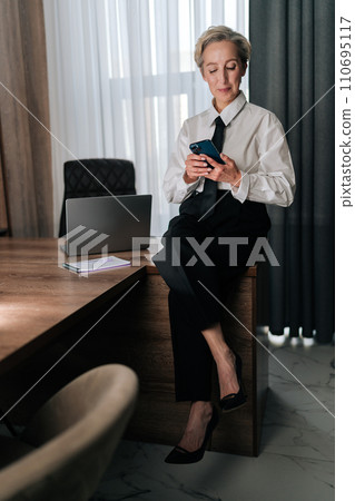 Vertical portrait of focused middle-aged business woman sitting on office table, looking at cellphone screen, making call by business, search information on internet distracted from work. Vertical portrait of focused middle-aged business woman sitting on office table, looking at cellphone screen, making call by business, search information on internet distracted from work. 110695117