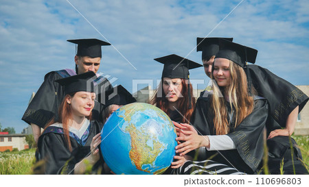 Graduates in black robes examine a geographical globe sitting on the grass. Graduates in black robes examine a geographical globe sitting on the grass. 110696803