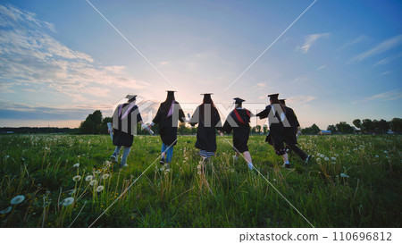 Six graduates in robes walk against the backdrop of the sunset. Six graduates in robes walk against the backdrop of the sunset. 110696812