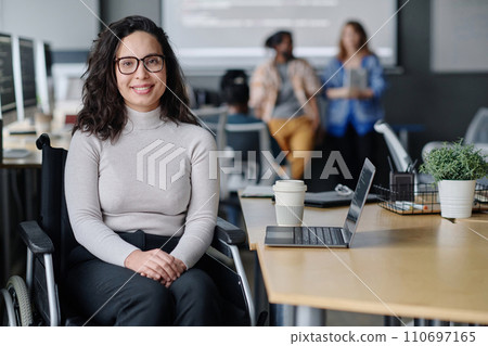 Smiling woman in wheelchair posing at workplace in office, her colleagues standing on background Smiling woman in wheelchair posing at workplace in office, her colleagues standing on background 110697165