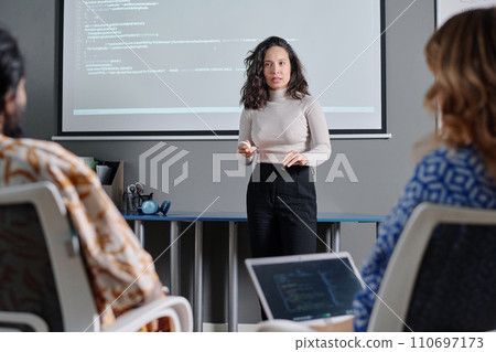 Serious female businesswoman talking to colleagues about code projected on digital board behind her Serious female businesswoman talking to colleagues about code projected on digital board behind her 110697173