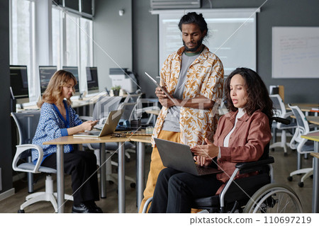 Woman with disability pointing at laptop screen while discussing project with her colleague Woman with disability pointing at laptop screen while discussing project with her colleague 110697219