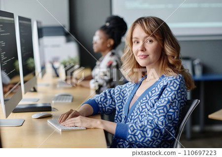 Caucasian female coder sitting at her desk working with data on computer and smiling at camera Caucasian female coder sitting at her desk working with data on computer and smiling at camera 110697223