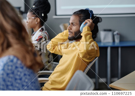 Indian office worker sitting at desk wearing headphones, his female colleagues working on background Indian office worker sitting at desk wearing headphones, his female colleagues working on background 110697225