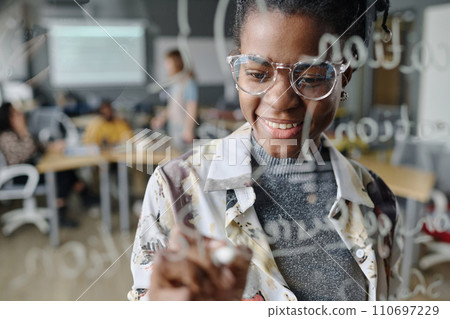 Cheerful woman making notes with marker on glass wall in office, her coworkers sitting on background 110697229