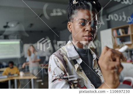 Female coder thoughtfully writing on glass wall in office, her colleagues working on background 110697230