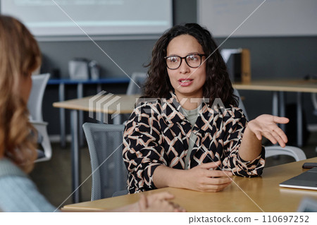 Young woman talking to her unrecognizable female colleague while sitting at desk in office 110697252