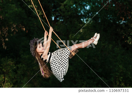 A small and beautiful girl in a dress is swinging on a swing. A small and beautiful girl in a dress is swinging on a swing. 110697331